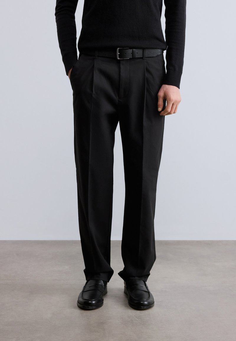 Μαύρο παντελόνι με άνετη εφαρμογή, με πιέτες μπροστά και ομαλή υφή, συνδυασμένο με μαύρη δερμάτινη ζώνη και παπούτσια.