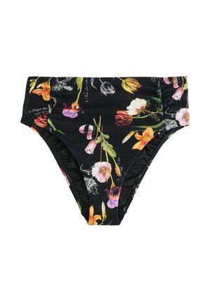 Femeie stă desculță pe țărmul stâncos purtând un costum de baie negru cu flori și cu spatele gol tip halter, cu oceanul și pământul îndepărtat sub un cer înnorat în fundal.