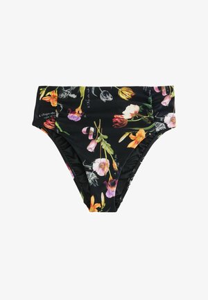 Frau steht barfuß am felsigen Ufer und trägt einen schwarzen, geblümten Neckholder-Bikini, im Hintergrund Meer und entferntes Land unter bewölktem Himmel.