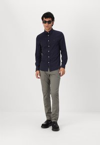 Donkerblauwe overhemden met lange mouwen, lichtgrijze chinos en zwarte schoenen. Model met zonnebril, staand tegen een effen achtergrond.