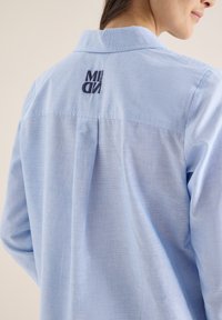 Femme portant une chemise bleu clair à col avec l'inscription "MIDN steady" en bleu foncé brodée sur le haut du dos, vue de dos et de profil.