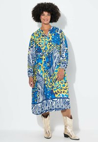 Kleurvolle maxi-jurk met een mix van blauw, geel en dierenprints. Het heeft lange mouwen, een V-hals en een strikdetail bij de taille. Gepaard met glanzende laarzen.