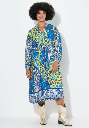 Abito maxi colorato con una combinazione di blu, giallo e stampe animali. Ha maniche lunghe, scollo a V e un dettaglio in vita con lacci. Abbinato a stivali lucidi.