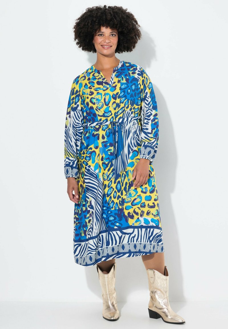 Kleurvolle maxi-jurk met een mix van blauw, geel en dierenprints. Het heeft lange mouwen, een V-hals en een strikdetail bij de taille. Gepaard met glanzende laarzen.