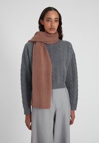Maglione grigio a trecce in maglia abbinato a una morbida sciarpa lunga e calda di colore marrone, avvolta attorno al collo. Pantaloni a gamba larga di un grigio chiaro completano l'outfit.