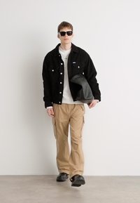 Black corduroy jacket, white sweatshirt, tan cargo pants, black sunglasses, and black leather bag, worn with black shoes; standing against a plain background.
