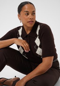Femme aux cheveux foncés tirés en arrière portant un pull marron à manches courtes avec un motif losanges blanc et un pantalon foncé, assise avec un genou relevé.