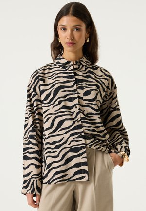 Femme aux cheveux longs porte une chemise ample à motif zébré et un pantalon beige, debout avec une main dans la poche devant un fond uni.