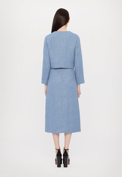 Blaues Tweedkleid mit einer kurzen Jacke, langen Ärmeln, geradem Schnitt und subtilen Glitzerdetails. Getragen mit schwarzen High Heels.