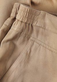 Beige stof met een gladde textuur, voorzien van een elastische tailleband en zijnaad. Over het algemeen een eenvoudig, praktisch ontwerp.