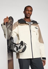 Casaco de esqui em creme e castanho com um capuz preto, apresentando um fecho de zip, um patch de logotipo, e emparelhado com uma prancha de snowboard e fixações.