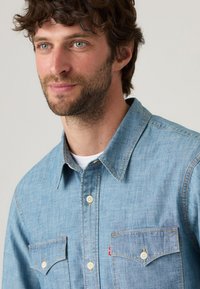 Light blue denim shirt featuring a button-down collar, two chest pockets with button closures, and contrast stitching details.