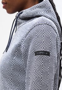 Προφίλ ατόμου που φοράει φούτερ με κουκούλα σε σχέδιο μπλε ναυτικό και λευκό με διαμάντι και λογότυπο Icepeak στο επάνω μέρος του βραχίονα.