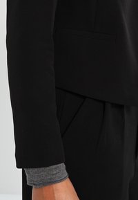 Černý blazer s přiléhavým střihem, hladkým materiálem a jednou náprsní kapsou. Šedé pletené manžety jsou viditelné pod rukávem blazera.