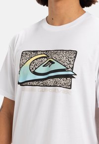 Camiseta blanca de algodón con un diseño colorido del logo de Quiksilver sobre un fondo negro texturizado y acentos en azul. Cuello redondo.