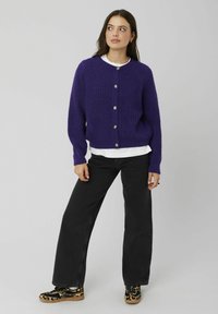 Cardigan violet en maille avec un col rond et une fermeture à boutons sur le devant, porté sur une chemise blanche et associé à un jean noir à jambes larges.