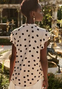 Blusa a pois in crema con maniche a sbuffo corte, caratterizzata da pois neri di varie dimensioni e un dettaglio di scollatura arricciata.
