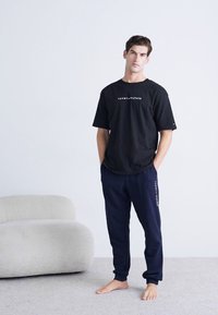 Schwarzes T-Shirt mit kurzen Ärmeln und "TOMMY HILFIGER"-Logo, kombiniert mit navy Sweatpants. Das Outfit hat eine lockere Passform und wird barfuß getragen.