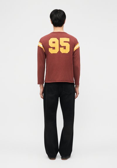 Homme debout, dos tourné, portant une chemise à manches longues bordeaux avec le numéro jaune "95" et des rayures sur les manches, associée à un pantalon noir.