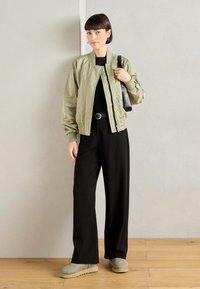 Chaqueta bomber verde oliva con puños de canalé, camiseta negra, pantalones anchos negros, botines verdes y un bolso negro sobre un fondo neutro.