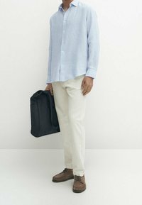 Camicia a righe blu chiaro con maniche lunghe, pantaloni beige, scarpe marroni. Borsa nera tenuta nella mano sinistra, su uno sfondo bianco.