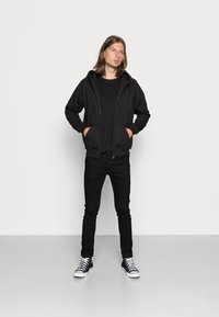 Sudadera negra con cremallera y bolsillos frontales, llevada sobre una camiseta negra. Combinada con jeans skinny negros y zapatillas altas negras con suelas blancas.