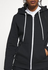 Felpa nera con zip realizzata in tessuto morbido. Presenta una tasca frontale, un cordino bianco e orlo a coste. Il cappuccio è funzionale e sagomato.