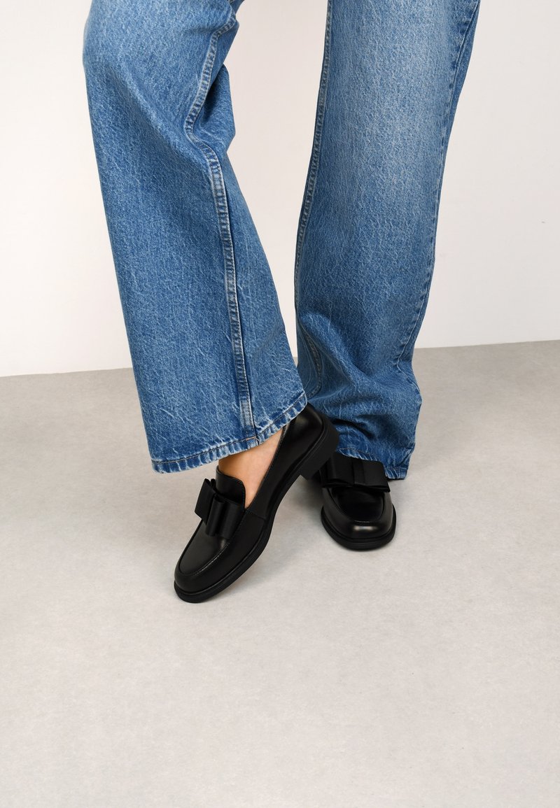 Black loafers with a bow detail, made of smooth leather. Rounded toe and cushioned sole. Paired with light blue wide-leg denim jeans.