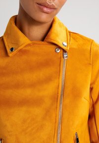 Gul mockaläder motorcykeljacka med knappkrage, silverdragkedja och sidofickor, som visar en jämn textur och en elegant design.