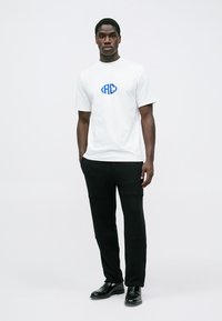 Man som står mot vit bakgrund iförd vit t-shirt med blå logotyp, svarta byxor och svarta finskor, med händerna i fickorna.
