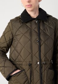 Gequilted olijfgroene jas met zwarte corduroy kraag, voorknopsluiting, trekkoord in de taille en twee zijzakken.