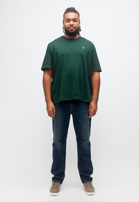 Άνδρας με πλεγμένα μαλλιά, ντυμένος με σκούρο πράσινο μπλουζάκι, μπλε τζιν και μπεζ αθλητικά παπούτσια, στέκεται μπροστά σε ένα απλό λευκό φόντο.