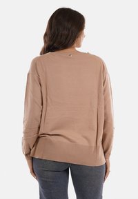 Donna con lunghi capelli castani che indossa un maglione beige largo e jeans blu scuro, in piedi con la schiena rivolta verso un sfondo semplice.