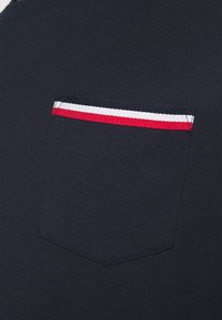 Mörkblå t-shirt med en texturerad ficka som har en röd, vit och blå randig detalj längs den övre kanten. Slät tyg.