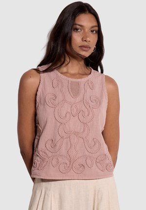 Mujer con cabello largo y oscuro que lleva una blusa sin mangas de color rosa con bordado elevado intrincado y pantalones plisados de color claro.