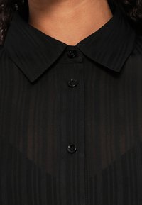 Melnā pogājama krekla ar apkakli kakla līnijā, kas izceļas ar smalkām vertikālām svītrām un melnām pogām priekšējā joslā.