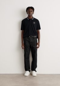 Zwarte polo met grijze kraagaccenten, gecombineerd met donkergrijze denim broek en witte sneakers. Het model staat tegen een lichte achtergrond.