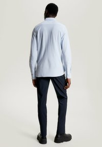 Camisa de rayas azul claro con mangas largas, puños abotonados y detalle de yugo en la parte posterior, combinada con pantalones oscuros y zapatos negros gruesos.