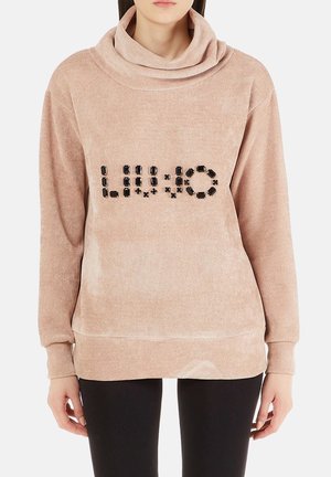 Femme portant un pull en velours côtelé beige avec un col haut, décoré de perles carrées noires formant un motif sur la poitrine.