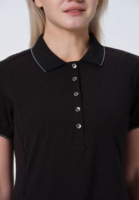 Polo noir avec un motif texturé en vagues, manches courtes, col noir avec des bordures blanches et boutons argentés sur le devant.
