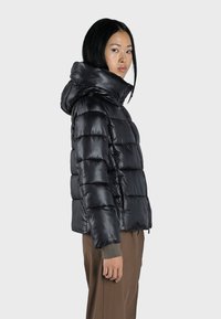 Zwarte pufferjack met capuchon, voorzien van een glanzende textuur, horizontale stiksels, zijzakken en elastische manchetten, gedragen over bruin broek.