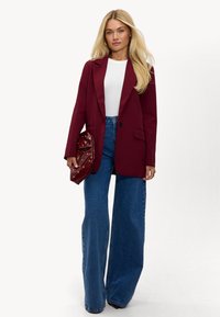 Bordeaux blazer met pieklappen, één knoopsluiting, gecombineerd met een aansluitend wit topje en wijdvallende blauwe denim jeans. Houdt een bordeaux rode clutch vast.