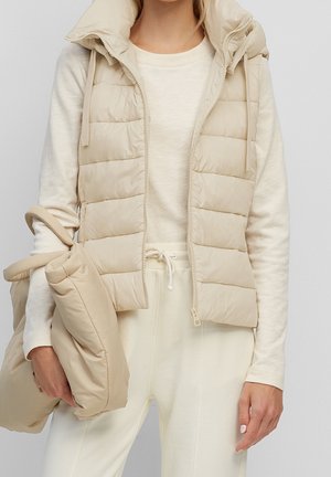 Vrouw draagt een beige doorgestikt vest over een crèmekleurig shirt met lange mouwen en crèmekleurige broek, en houdt een bijpassende beige doorgestikte tas vast.