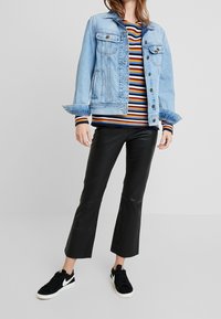 Veste en jean bleu clair au design classique, portée sur une chemise rayée avec des lignes rouges, jaunes et bleues, associée à un pantalon en fausse cuir noir et des baskets noires.