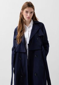 Cabană navy dintr-un material neted, cu rever mari, design cu două rânduri de nasturi și buzunare laterale, îmbrăcată cu o cămașă albă cu guler.