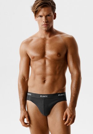 Wytrenowany mężczyzna o jasnobrązowych włosach, ubrany w czarne slipy marki Atlantic, stoi na jednolitym jasnym tle, zwrócony twarzą do przodu.