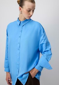 Egy kontyba tűzött hajjal rendelkező nő bő szabású, élénk kék inget visel, az egyik kezét a zsebében tartja, és lefelé, balra néz.