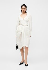 Nő fehér, hosszú ujjú öves ruhában, drapírozott részlettel, kezében egy kis fekete kézitáskával és fekete hegyes orrú magassarkú cipőkkel, fehér háttér előtt áll.