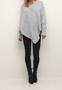 Kvinna i en grå stickad poncho med zigzagmönster, svarta tajta byxor och svarta ankelboots som står mot en enkel ljus bakgrund.