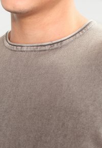 T-shirt en coton brun clair avec un col rond et une texture côtelée. Détails de couture visibles le long du col et des coutures d'épaule.